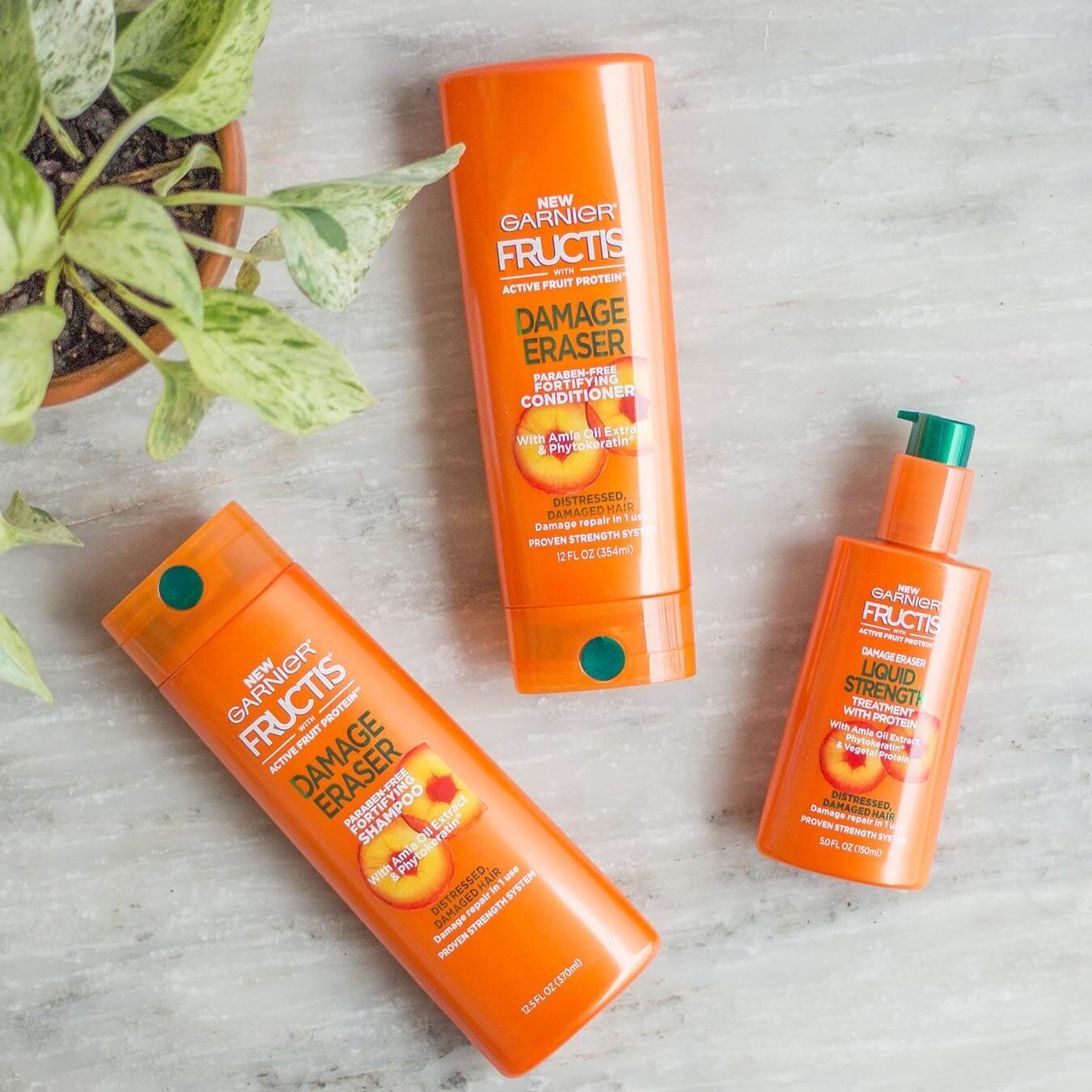 Garnier Fructis Damage Eraser Shampoo, Damage Eraser Conditioner, and Fructis Liquid Strength Treatment on gray marble next to a potted Chinese Evergreen houseplant.